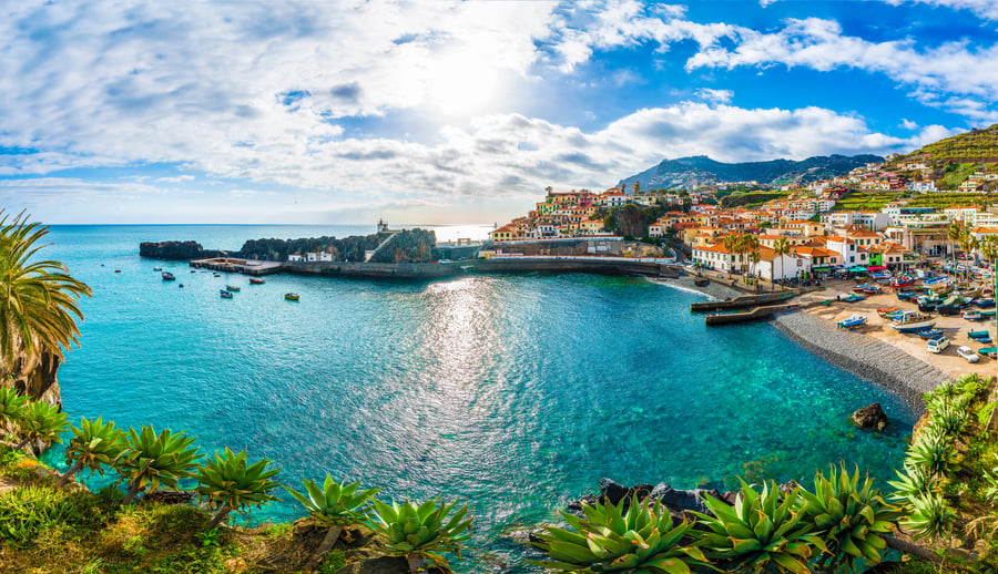 Panoramic view of Câmara de Lobos coastal village in Madeira, with turquoise Atlantic waters, fishing boats, and colorful houses under the sun.