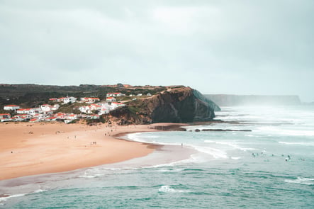 Aerial view of Arrifana Beach on the Vicentine Coast, with dramatic cliffs, surfers in the sea, and traditional white Algarve houses.