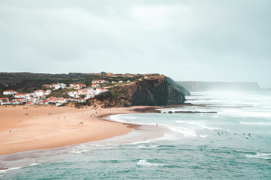 Aerial view of Arrifana Beach on the Vicentine Coast, with dramatic cliffs, surfers in the sea, and traditional white Algarve houses.
