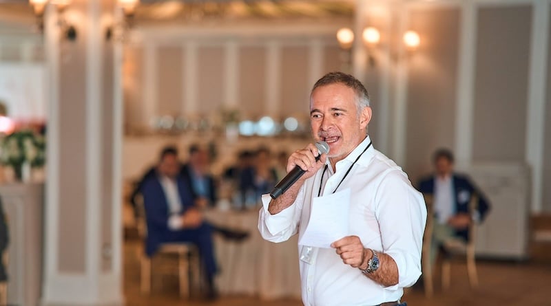 Man speaking into a microphone during a corporate event, with seated attendees in an elegant conference room.
