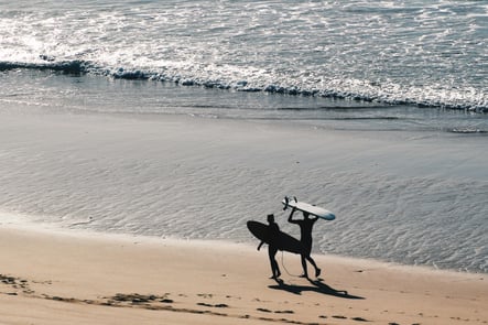 Two surfers walking along Sagres beach at sunset, carrying surfboards by the Atlantic Ocean waves.