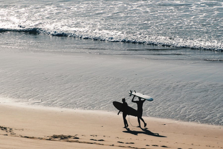 Two surfers walking along Sagres beach at sunset, carrying surfboards by the Atlantic Ocean waves.