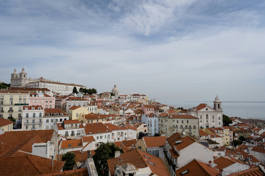 Panoramic view of Alfama district in Lisbon, with red rooftops and the Tagus River in the background.