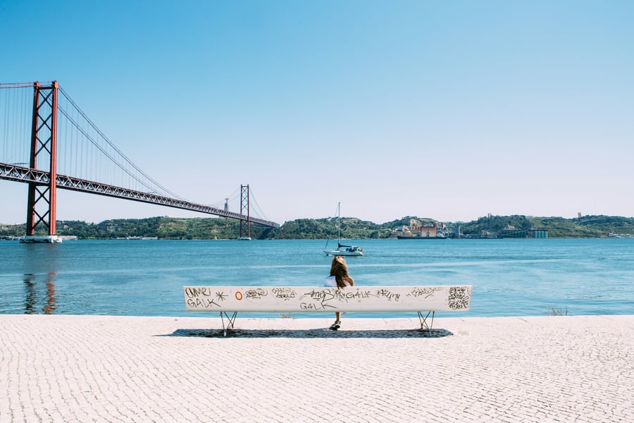 View of the 25 de Abril Bridge in Lisbon with a woman sitting by the Tagus River under a blue sky, representing Portuguese lifestyle and serenity.