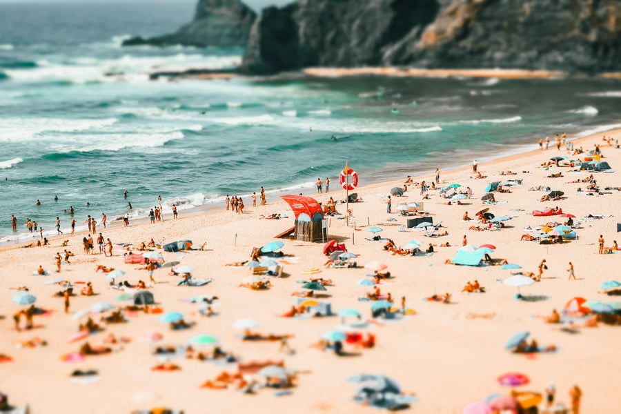 Aerial view of a lively beach on the Portuguese coast, with sunbathers, colorful umbrellas, and clear blue sea during summer.