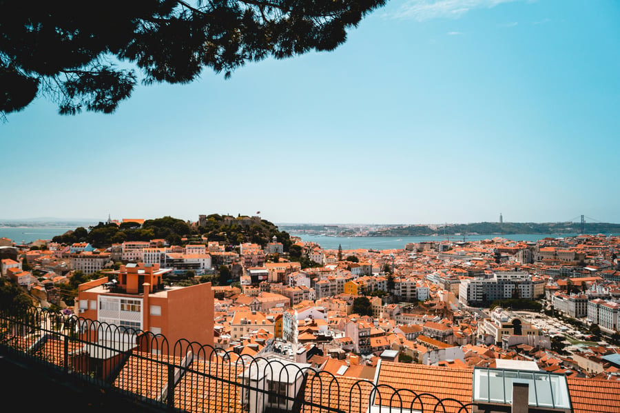 Panoramic view of Lisbon featuring São Jorge Castle, the Tagus River, and the city’s iconic orange rooftops under a clear blue sky.