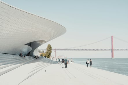 Modern view of the MAAT in Lisbon, Portugal, with the 25 de Abril Bridge in the background and people walking along the Tagus River on a sunny day.