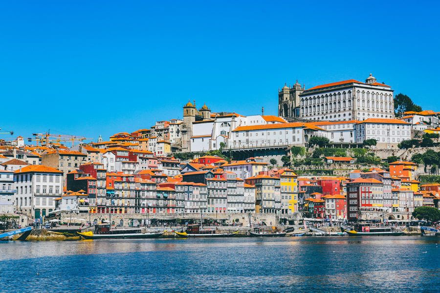 Panoramic view of Ribeira in Porto, Portugal, featuring colorful riverside buildings along the Douro River and the Serra do Pilar Monastery under a bright blue sky.