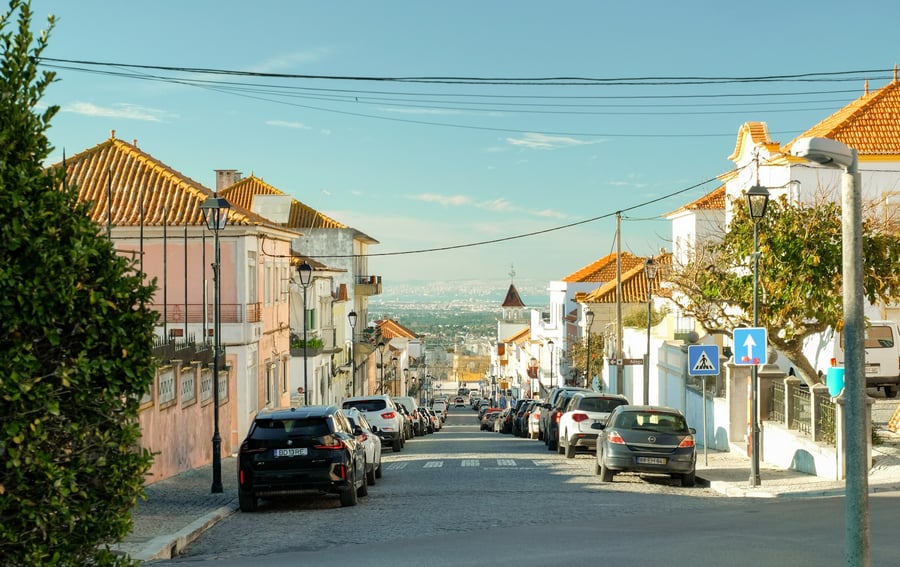 View of a typical street in Almada, Portugal, featuring orange-tiled houses, parked cars, and the Tagus River in the background — an authentic scene that captures the charm of Portuguese towns near Lisbon.