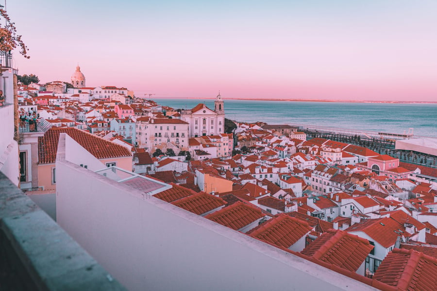 Panoramic view of Alfama at sunset, with red rooftops, the Tagus River, and traditional Lisbon architecture glowing in pink tones.