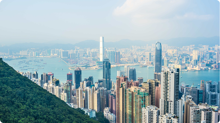 Panoramic view of Hong Kong with modern skyscrapers, the harbor in the background, and lush green mountains.