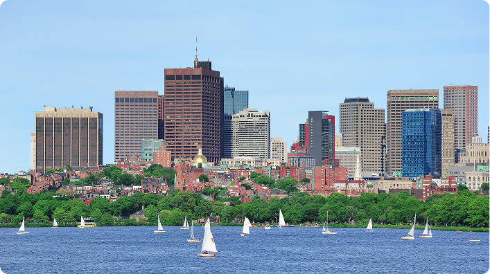 Panoramic view of Boston with sailboats on the Charles River and skyscrapers in the background.
