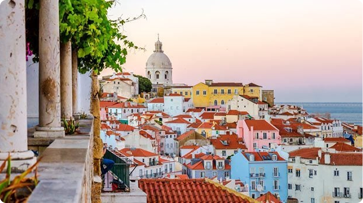 Vista do bairro de Alfama em Lisboa ao pôr do sol, com casas coloridas, telhados de terracota e o Panteão Nacional ao fundo.